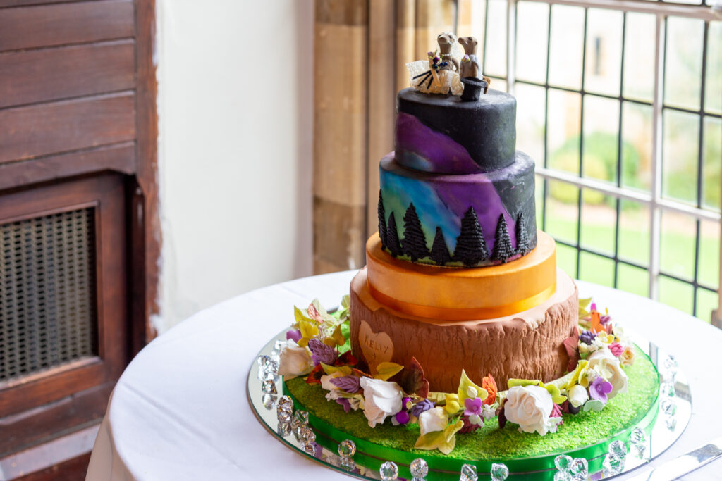 Photograph of wedding cake with 3 tiers. The cake has a aurora and autumn theme with otters getting married as a topper